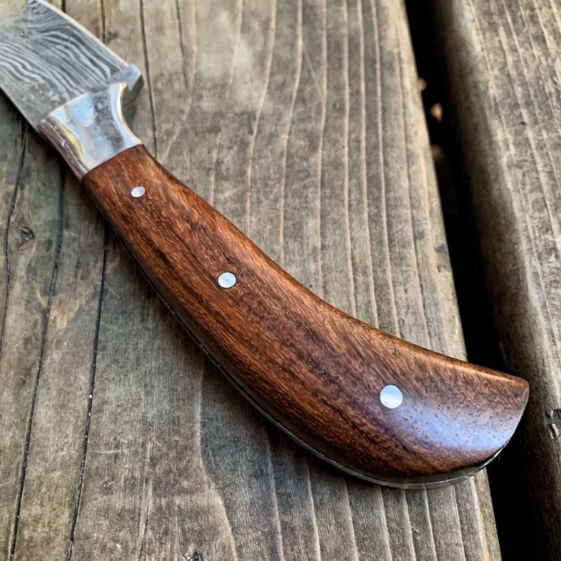 May include: Close-up of a Damascus steel knife with a dark brown wooden handle. The handle is adorned with three silver rivets. The blade has a silver bolster and a patterned design.