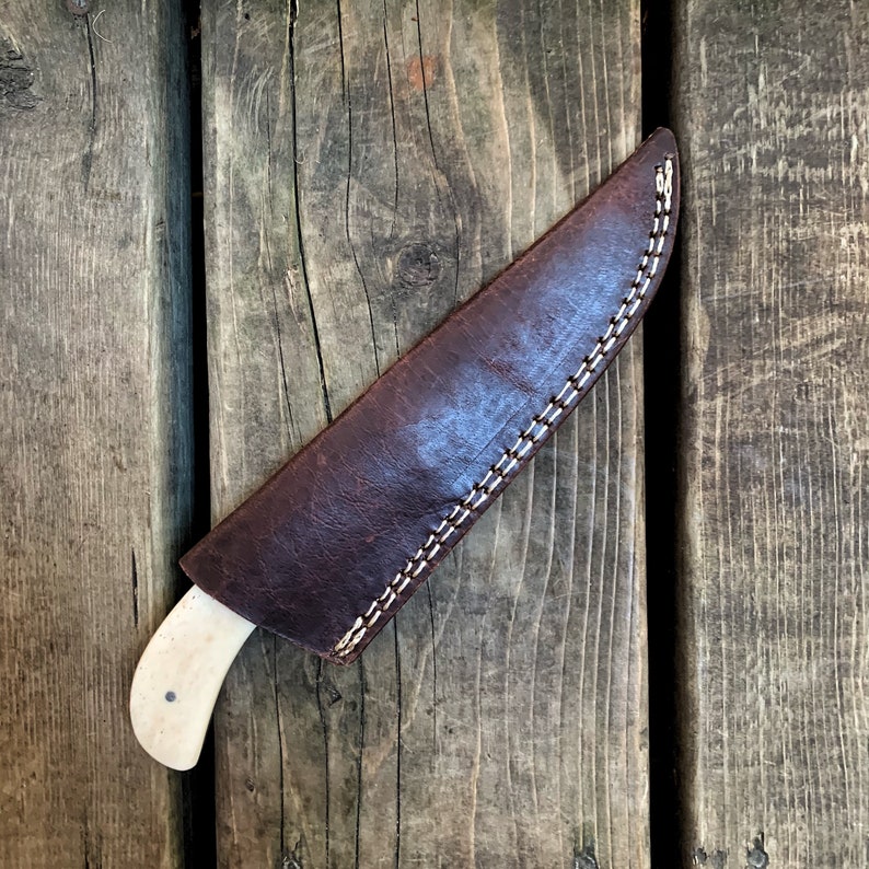 May include: A handcrafted knife with a bone handle is partially sheathed in a dark brown leather case. The case is stitched with white thread. The knife and sheath rest on a weathered wooden surface.