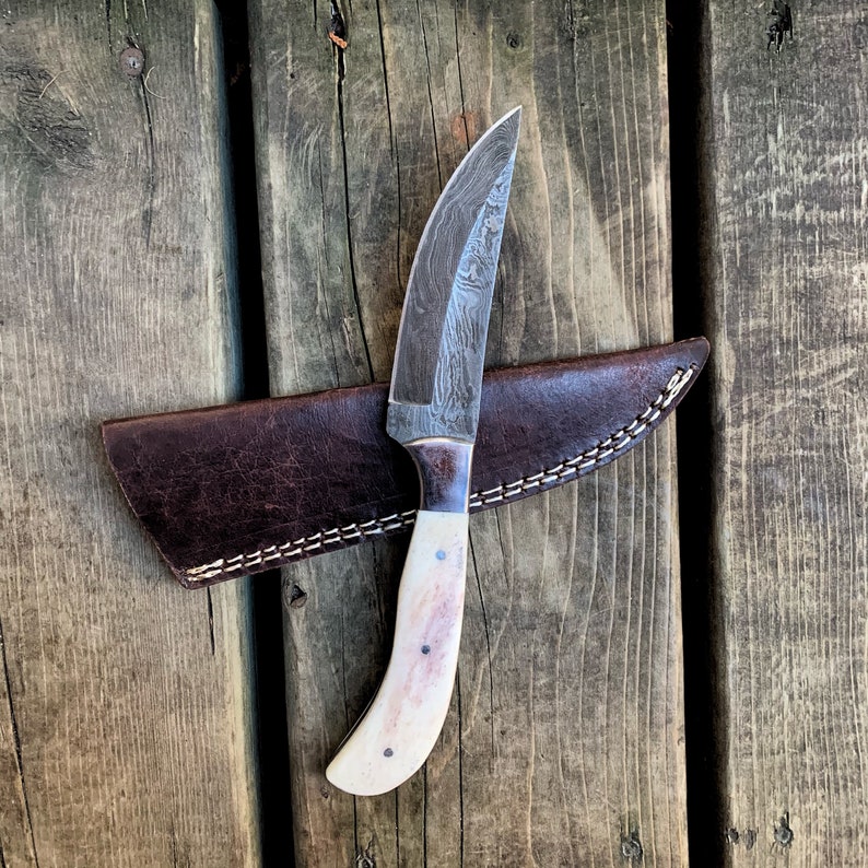 May include: A handcrafted hunting knife with a patterned steel blade and a bone handle. The knife rests on a brown leather sheath with white stitching, set against a wooden background. The blade is approximately 7 inches long.