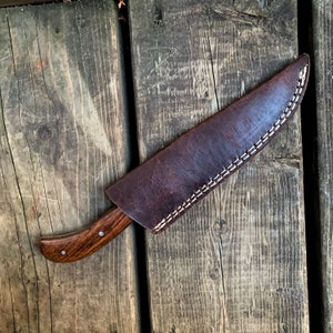 May include: A handcrafted hunting knife with a dark wooden handle and a brown leather sheath. The blade is partially visible, and the sheath is stitched with white thread. The knife rests on a weathered wooden surface.