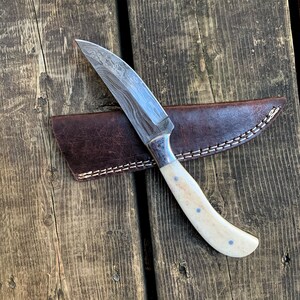 May include: A handcrafted hunting knife with a patterned steel blade and a bone handle with blue dot accents. The knife rests on a wooden surface next to a brown leather sheath with white stitching.