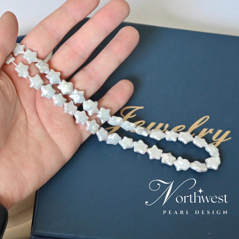 May include: A strand of white pearl beads shaped like stars. The beads are strung on a white thread and are displayed on a hand against a blue background. The background has the text "Jewelry Northwest Pearl Design".