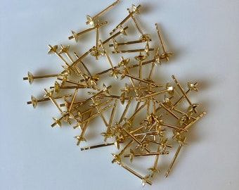 May include: A pile of gold-toned earring posts, also known as earring findings, on a white surface.