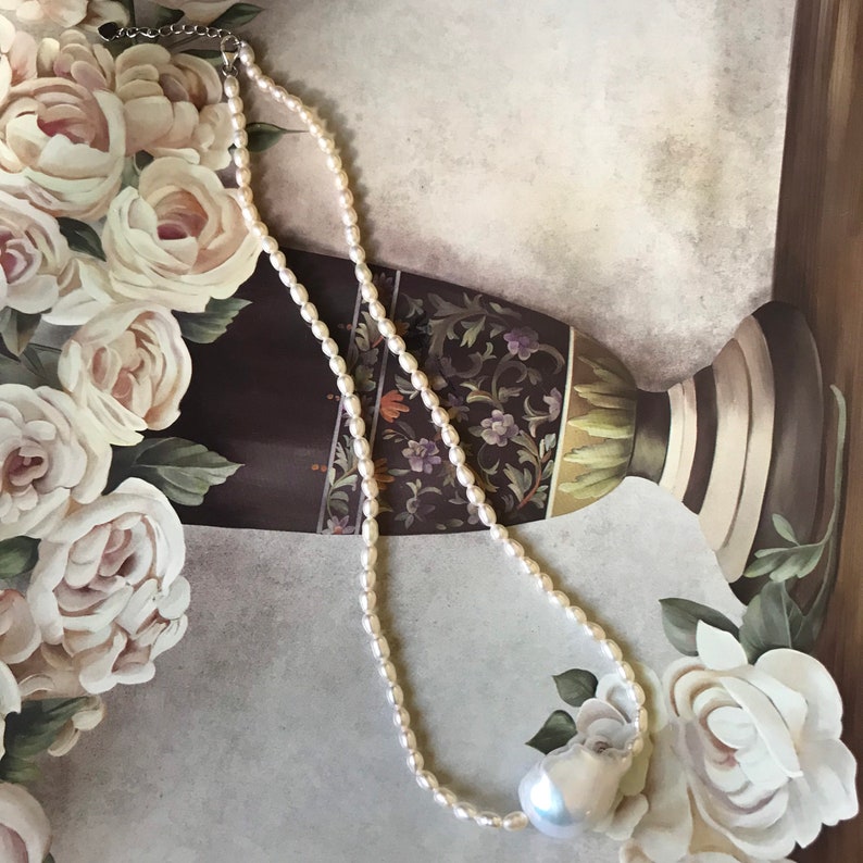 Peut inclure: Un collier de perles blanches avec un grand pendentif en perle blanc cass&eacute;. Le collier est pos&eacute; sur une surface &agrave; motifs floraux.