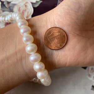 May include: A white pearl bracelet with a single strand of pearls. The bracelet is shown on a person's wrist next to a US penny.
