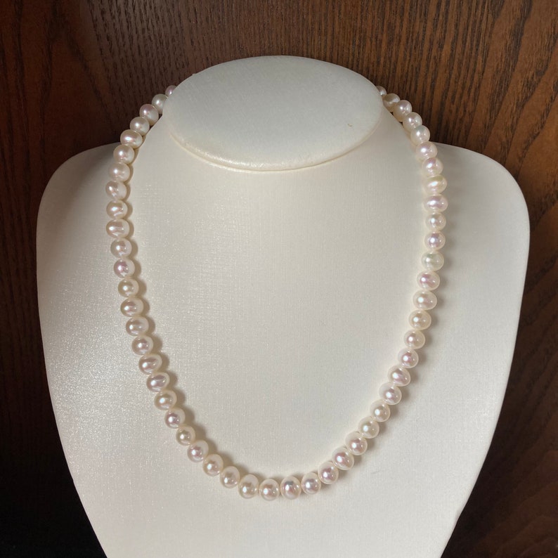 May include: A strand of white pearl necklace on a white display stand. The pearls are round and smooth.