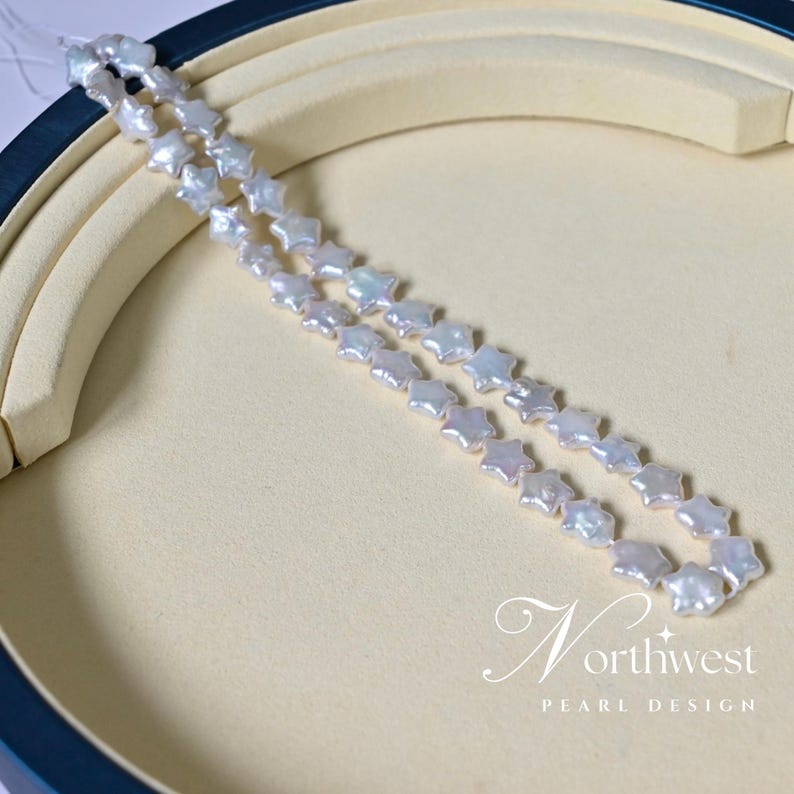 May include: A strand of white freshwater pearls in a star shape, displayed on a white background. The pearls are strung together on a clear thread. The pearls are iridescent and have a slight pink hue. The image includes the text "Northwest Pearl Design".