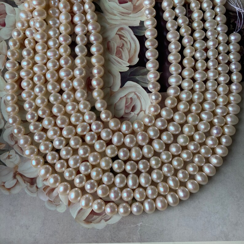 May include: A strand of white freshwater pearls, with a smooth, round shape. The pearls are strung on a clear thread and are arranged in a loose, flowing pattern.