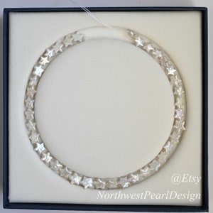 May include: A white pearl necklace with star-shaped beads in a white box. The necklace is in a circle shape and the box has the text "@Etsy NorthwestPearlDesign" on the bottom right corner.