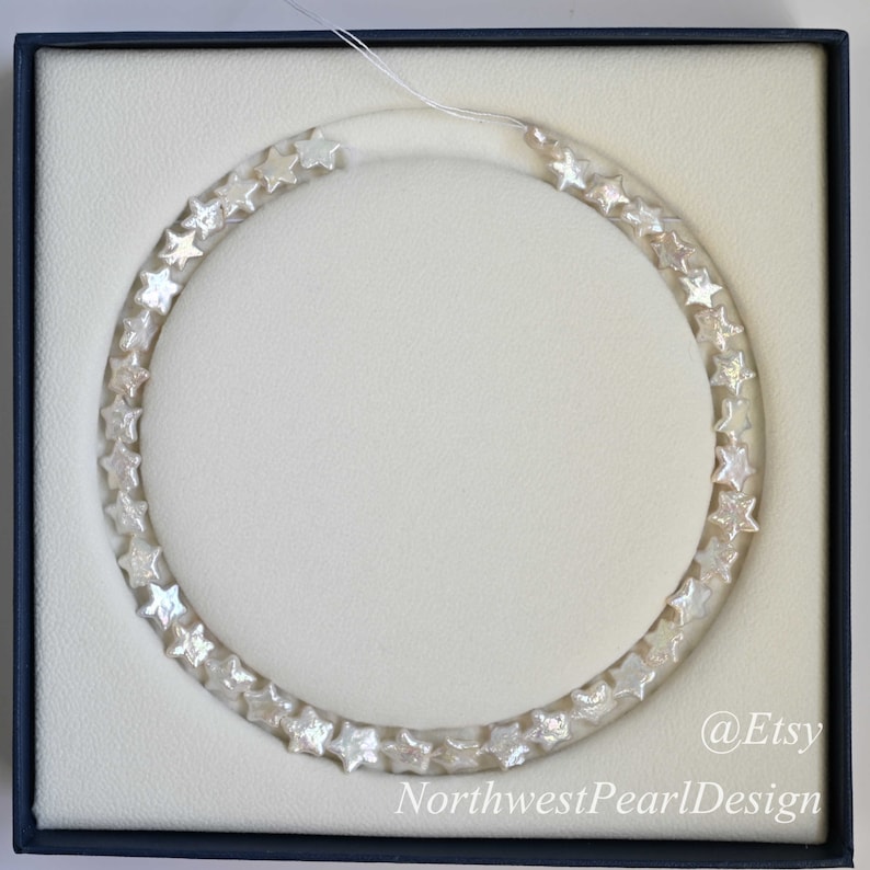 May include: A white pearl necklace with star-shaped beads in a white box. The necklace is in a circle shape and the box has the text "@Etsy NorthwestPearlDesign" on the bottom right corner.