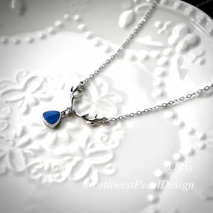 Op de afbeelding: Een zilveren ketting met een blauwe opaal hanger en een zilveren hertengewei ontwerp. De ketting ligt op een wit oppervlak met een bloemenpatroon.