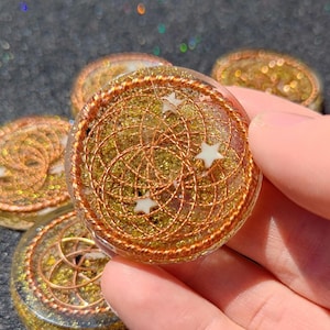 May include: A circular resin disc containing gold-colored metallic flakes and a coiled copper wire pattern arranged in overlapping circles. Three small, white, five-pointed star-shaped accents are embedded within the clear, polished resin surface.