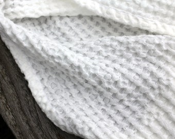 May include: Close-up of a white waffle-weave fabric. The fabric has a textured, honeycomb-like pattern. The material is draped, with soft folds and shadows. The background is a weathered wooden surface.