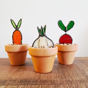 May include: Three small terracotta pots each hold a different stained glass vegetable: a carrot, an onion, and a radish. The carrot is orange with green leaves, the onion is white with green leaves, and the radish is red with green leaves.