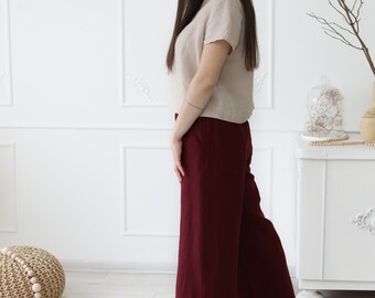 May include: A woman wearing a beige linen top and wide-legged burgundy linen pants. She is standing on a round jute rug with a woven ball and string of beads in the background.