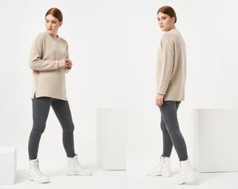 May include: A beige sweater with long sleeves and a relaxed fit. The sweater has a round neckline and a slightly longer length in the back. The model is wearing dark gray skinny jeans and white ankle boots.