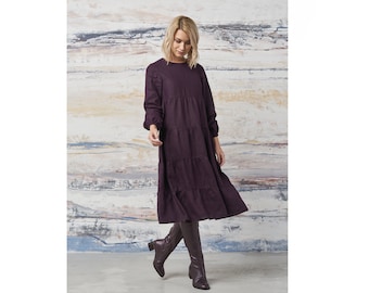 May include: A long-sleeved, tiered dress in a deep purple color. The dress has a round neckline and a relaxed fit. The model is wearing the dress with dark brown boots.