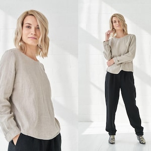 Può includere: Due donne indossano una blusa di lino beige chiaro a maniche lunghe. La blusa ha una scollatura rotonda e una vestibilità rilassata. Le donne indossano pantaloni neri.