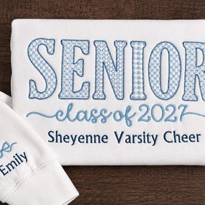 May include: A white sweatshirt featuring blue embroidered text that reads SENIOR class of 2027 Sheyenne Varsity Cheer. The sleeve cuff displays the name Emily in blue embroidery above a small blue bow icon. The embroidery has a checkered pattern.