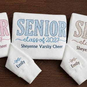 May include: Three white sweatshirts featuring embroidered text. Each displays SENIOR class of 2027 Sheyenne Varsity Cheer in pink, blue, or brown. The sleeves are folded to show the name Emily embroidered in matching colors with a small bow icon.