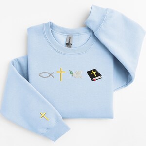 May include: A light blue sweatshirt with a Christian symbol design. The symbols include a fish, a cross, an olive branch, a dove, and a closed book with a cross on the cover.