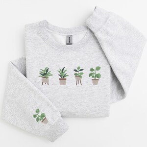 Pode incluir: Uma camisa de moletom cinza com um gr&aacute;fico de cinco plantas em vasos. As plantas s&atilde;o verdes e marrons e est&atilde;o dispostas em uma fileira na frente da camisa de moletom.