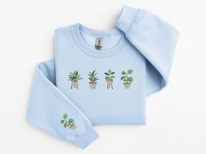 Pode incluir: Uma camisola azul claro com um gr&aacute;fico de cinco plantas em vasos bordados na frente. A camisola tem um decote redondo e mangas compridas.