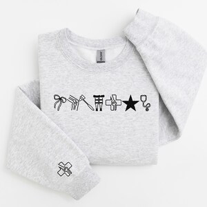 May include: A light gray sweatshirt with black line art illustrations of medical symbols, including a bow, knee, hammer, and star. The sleeve cuff features a cross symbol. The sweatshirt is folded.