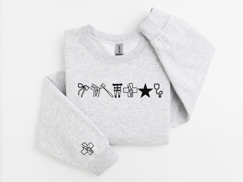 May include: A light gray sweatshirt with black line art illustrations of medical symbols, including a bow, knee, hammer, and star. The sleeve cuff features a cross symbol. The sweatshirt is folded.