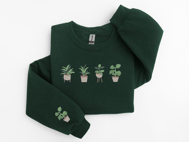Pode incluir: Uma camisola verde escura com imagens bordadas de plantas em vasos. As plantas s&atilde;o de estilos e cores diferentes. A camisola tem gola redonda e mangas compridas.