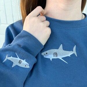 May include: Indigo blue sweatshirt with a white embroidered shark on the front and a smaller shark on the sleeve. "Indigo Blue is Color Shown" is written at the bottom of the image.