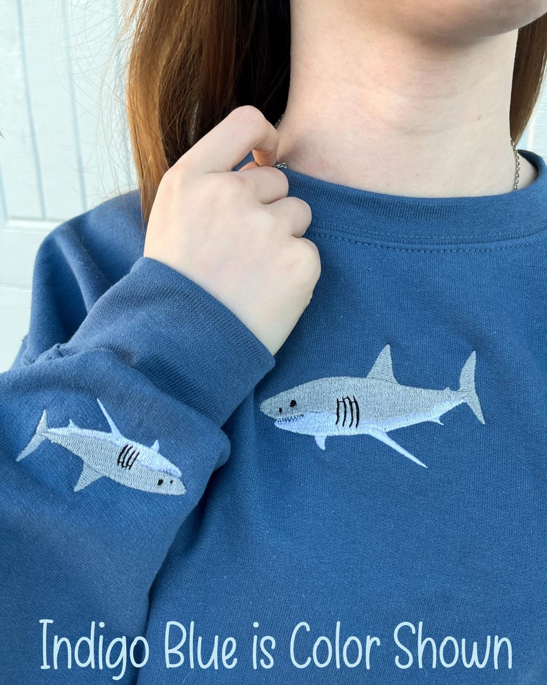 May include: Indigo blue sweatshirt with a white embroidered shark on the front and a smaller shark on the sleeve. "Indigo Blue is Color Shown" is written at the bottom of the image.