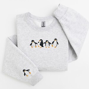 Pu&ograve; includere: Una felpa grigio chiaro con girocollo e maniche lunghe. La parte anteriore della felpa presenta un disegno ricamato di cinque pinguini. Un pinguino &egrave; ricamato sulla manica sinistra.