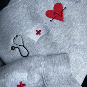 May include: Gray sweatshirt with embroidered medical symbols, including a stethoscope, a heart with a heartbeat line, a syringe, and a red cross in a white square. The sweatshirt has a red cross in a white square on the sleeve.