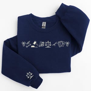 Pode incluir: Camisola azul marinho com desenhos a tra&ccedil;o brancos de s&iacute;mbolos legais e texto. O design inclui um martelo, balan&ccedil;as e documentos. A manga apresenta um &iacute;cone de balan&ccedil;as. O texto inclui "Coffee break" e "Urgent!"