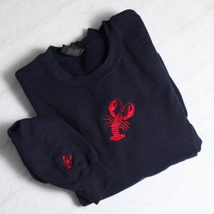 May include: A navy blue sweatshirt with a red embroidered lobster on the front. The sweatshirt has a small red embroidered lobster on the sleeve.
