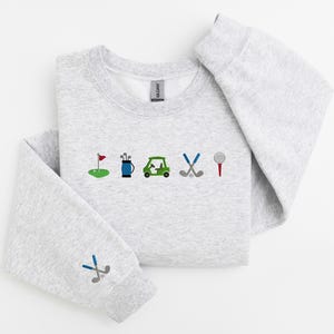 以下が含まれることがあります： グレーのトレーナーに、前面にゴルフをテーマにした刺繍が施されています。デザインには、ゴルフの旗、ゴルフバッグ、ゴルフカート、ゴルフクラブ、ゴルフボールが含まれています。