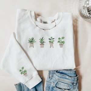 Pode incluir: Camisola branca com um gr&aacute;fico de cinco plantas em vasos. As plantas s&atilde;o de estilos diferentes e t&ecirc;m folhas verdes. A camisola est&aacute; sobre uma superf&iacute;cie castanho claro.