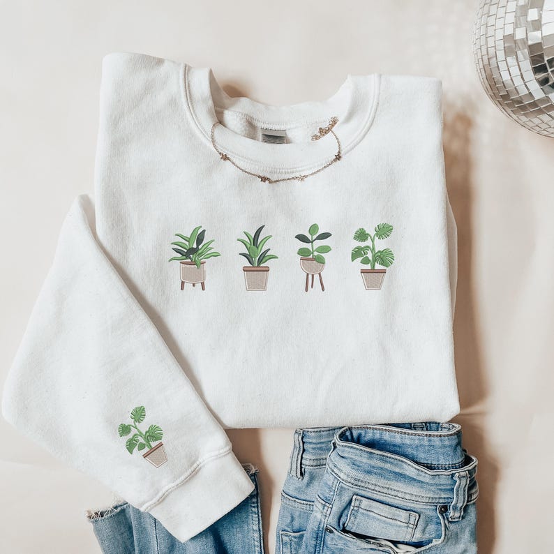 Pode incluir: Camisola branca com um gr&aacute;fico de cinco plantas em vasos. As plantas s&atilde;o de estilos diferentes e t&ecirc;m folhas verdes. A camisola est&aacute; sobre uma superf&iacute;cie castanho claro.