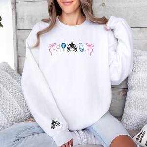 May include: White sweatshirt with embroidered medical-themed designs. Features pink bows, a stethoscope, lungs, and a stethoscope in blue. A lung design is embroidered on the sleeve. The sweatshirt is worn by a person.