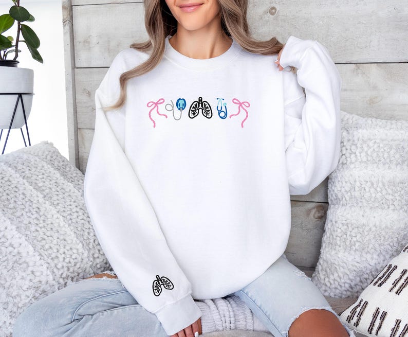 May include: White sweatshirt with embroidered medical-themed designs. Features pink bows, a stethoscope, lungs, and a stethoscope in blue. A lung design is embroidered on the sleeve. The sweatshirt is worn by a person.