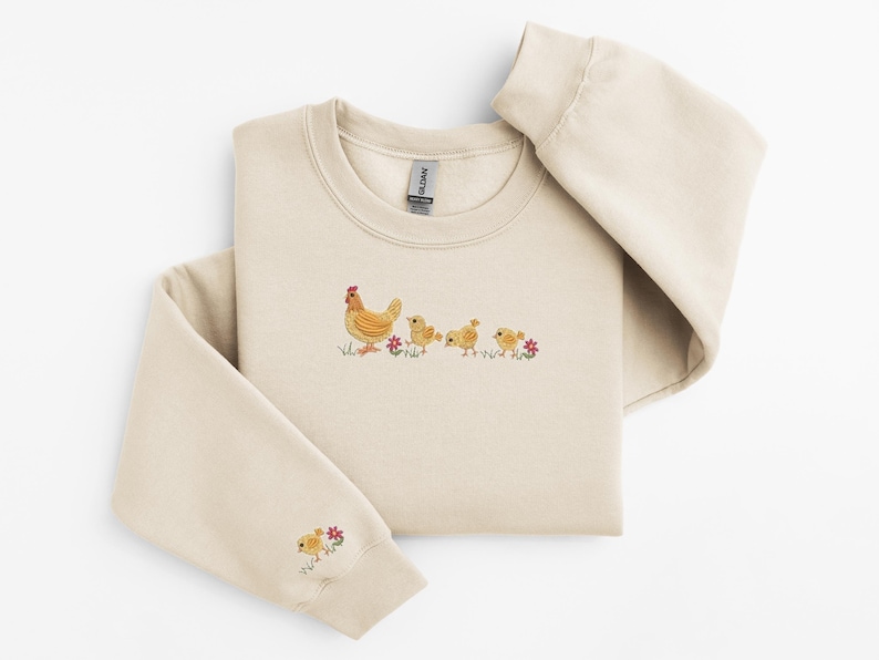 K&ouml;nnte beinhalten: Ein beiger Pullover mit einem gestickten Design einer Henne mit ihren K&uuml;ken. Das Design ist in Gelb- und Braunt&ouml;nen mit roten Blumen gehalten.