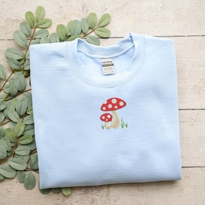 Pode incluir: Uma camisa de frio azul claro com uma imagem bordada de dois cogumelos vermelhos e brancos com hastes marrons. Os cogumelos s&atilde;o cercados por grama verde.