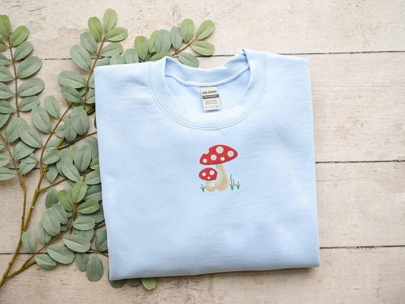 Pode incluir: Uma camisa de frio azul claro com uma imagem bordada de dois cogumelos vermelhos e brancos com hastes marrons. Os cogumelos s&atilde;o cercados por grama verde.