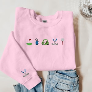 以下が含まれることがあります： ゴルフをテーマにした刺繍デザインが施されたピンク色のスウェットシャツ。デザインには、ゴルフカート、ゴルフクラブ、ゴルフバッグ、ゴルフボール、旗が含まれています。