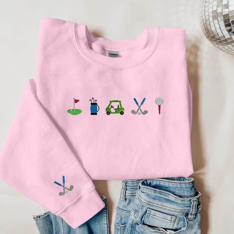 以下が含まれることがあります： ゴルフをテーマにした刺繍デザインが施されたピンク色のスウェットシャツ。デザインには、ゴルフカート、ゴルフクラブ、ゴルフバッグ、ゴルフボール、旗が含まれています。