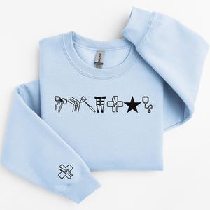 May include: A light blue sweatshirt with black medical-themed graphics. The design includes a bow, knee, hammer, surgical tools, cross, star, and stethoscope. A matching graphic is on the sleeve.