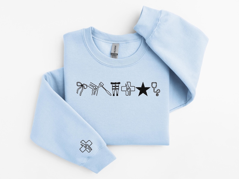 May include: A light blue sweatshirt with black medical-themed graphics. The design includes a bow, knee, hammer, surgical tools, cross, star, and stethoscope. A matching graphic is on the sleeve.