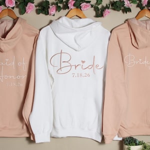 May include: Three hooded sweatshirts in blush pink and white with the words "Maid of Honor", "Bride", and "Bridesmaid" in cursive script with a small heart and the date "7.18.26" printed on the back.