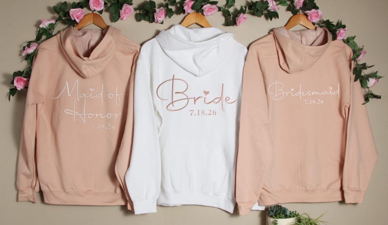 Pu&ograve; includere: Tre felpe con cappuccio in rosa cipria e bianco con le parole "Maid of Honor", "Bride" e "Bridesmaid" in corsivo con un piccolo cuore e la data "7.18.26" stampata sul retro.
